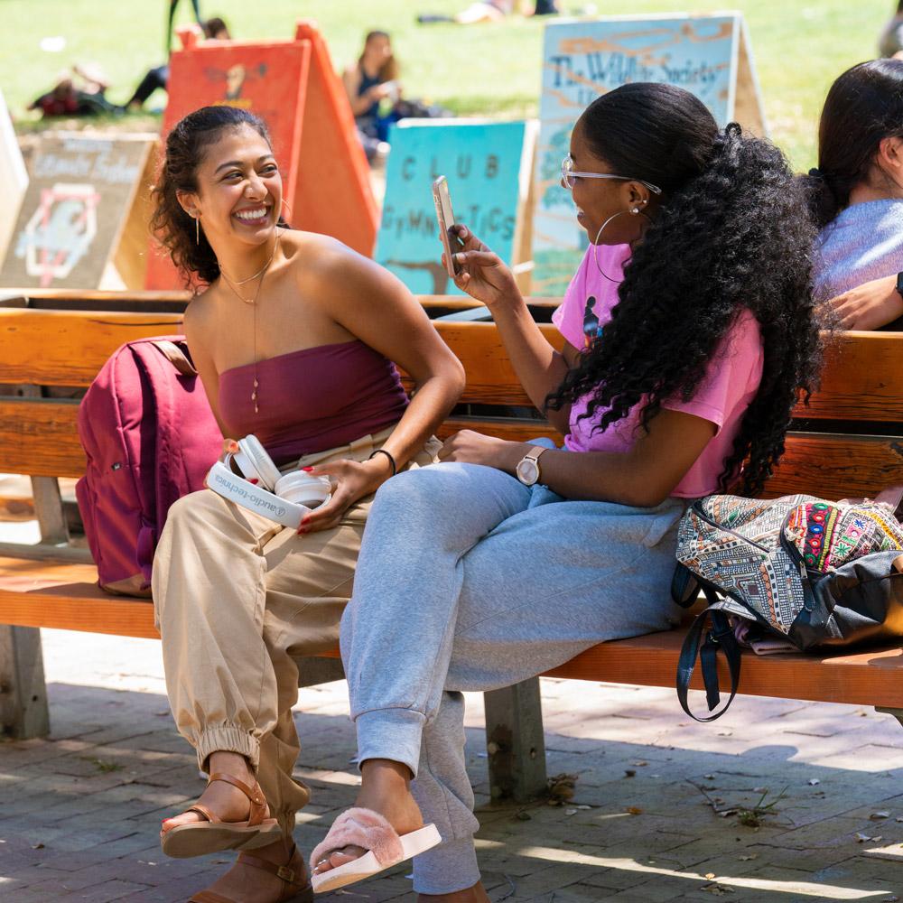 two students sitting on a bench near the quad with signs of student organizations in the background