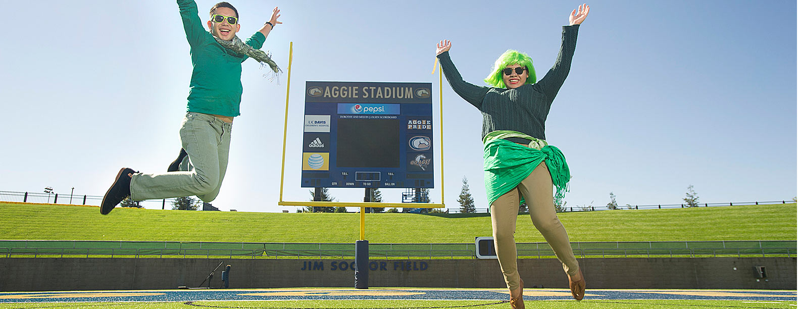A woman and a man dressed in several pieces of green clothing pose in front of the Aggie Stadium scoreboard