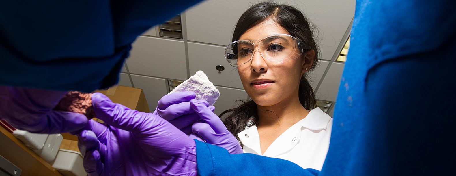 Woman in lab coat holds a rock while another pair of hands holds another rockin the foreground