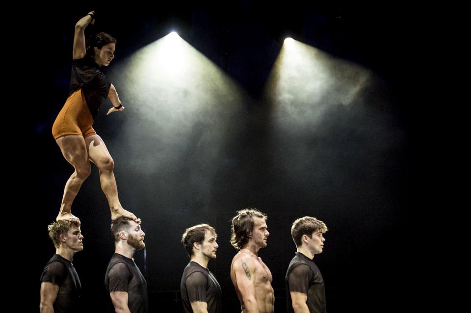 Circa performing on stage with five men standing in line and a woman walking on the heads of two of them.