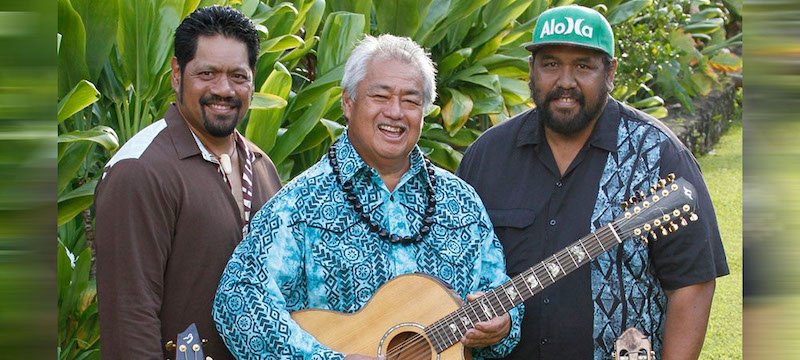 George Kahumoku Jr., Nathan Aweau and David “Kawika” Kahiapo standing in front of green Hawaiian foliage.